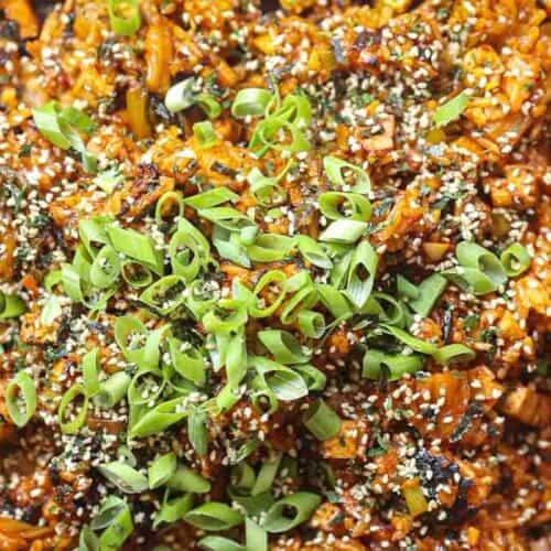 Close-up of a vibrant dish featuring vegan kimchi fried rice mixed with chopped vegetables. The dish is topped with a generous amount of sliced green onions and sprinkled with sesame seeds, giving it a colorful and textured appearance.